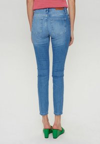 Gai zilu šauras džinsu bikses ar gludu denimu, augstu jostasvietu, piecām kabatām un kontrastējošām šuvēm. Valkātas ar zaļiem platformas papēžiem.