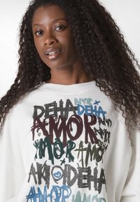 Sudadera blanca con texto variado en negro, verde, azul y burdeos, con una combinación de acabados mate y brillante.