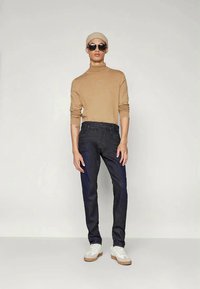 Pullover de gola alta bege, jeans escuros, ténis brancos. A modelo usa óculos de sol e um gorro combinando. Textura suave, design ajustado, estilo casual.