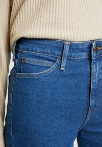 Jean en denim bleu avec une taille mi-haute, design à cinq poches et surpiqûres orange contrastantes. Associé à un pull beige texturé.