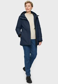 Vrouw die wandelt in een marineblauwe winterjas met capuchon, een beige gebreid vest, blauwe jeans en zwarte laarzen op een witte achtergrond.