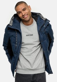 Marineblauwe pufferjack met synthetisch materiaal en zwarte accenten, voorzien van een ritssluiting en capuchon. Daaronder een lichtgrijs sweatshirt met bedrukte logo.