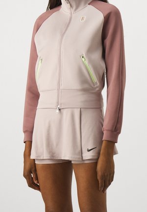 Kvinde iført lyserød lynlåstrøje med mavefarvede ærmer og matchende plisseret nederdel med sort Nike-logo på venstre kant.