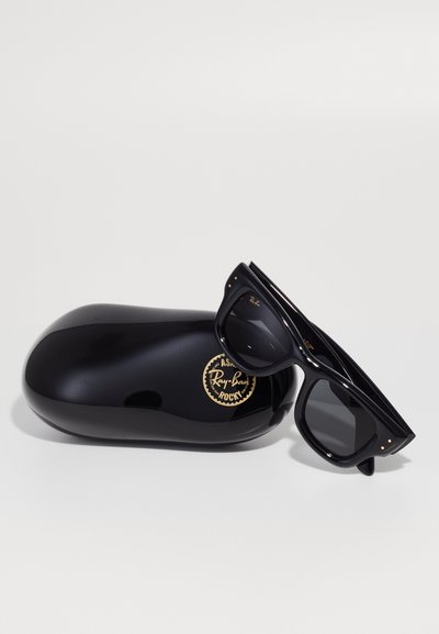 Черни слънчеви очила Ray-Ban с тъмни лещи са разположени върху лъскава черна кутия, украсена с златист кръгъл лого. Очилата имат гладки рамки.