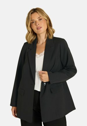 Frau mit blonden Haaren, die einen schwarzen Blazer über einem weißen, mit Spitze verzierten Top trägt und mit einer Hand den Blazer in der Nähe des Knopfs hält.