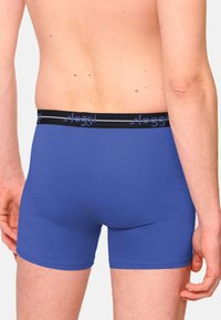 Blaue Boxershorts mit einem schwarzen elastischen Bund, der ein Logo trägt. Glatter Stoff mit enger Passform und minimalen Nähten.
