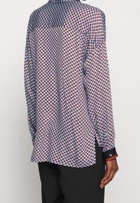 Long-sleeve shirt in lightweight fabric featuring a blue base with red polka dots, with contrasting cuffs and side slits for mobility.