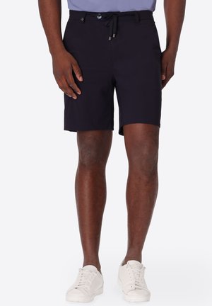 Uomo che indossa pantaloncini neri fino al ginocchio con bottone e coulisse, abbinati a sneakers bianche, in piedi su uno sfondo semplice.