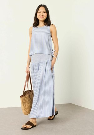 Junge Frau, die drinnen geht, trägt ein blau-weiß gestreiftes ärmelloses Top und einen langen Rock, trägt eine geflochtene Tasche und braune Sandalen.