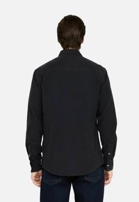 Uomo con capelli castani che indossa una camicia nera a maniche lunghe con bottoni e jeans blu scuro, girato di spalle su uno sfondo chiaro e semplice.