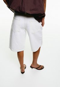 Bílé džínové šortky se uvolněným střihem a dvěma zadními kapsami, doplněné hnědými žabkami, pohled zezadu.