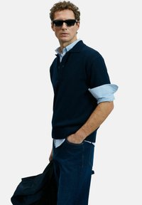 Mann mit schwarzen Sonnenbrille, navyfarbener Pullover über hochgekrempeltem gestreiften Hemd, blauen Jeans, hält eine navyfarbene Jacke mit einer Hand in der Tasche.