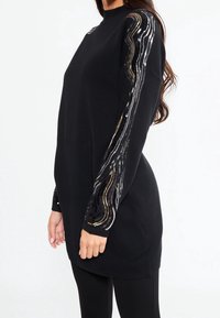 Donna che indossa un lungo maglione nero con strisce di paillettes argentate e dorate che decorrono lungo le maniche, abbinato a leggings neri.