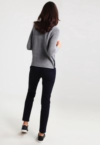 Femme aux longs cheveux bruns portant un pull gris, un pantalon noir et des baskets noires, debout et vue de dos sur un fond blanc.