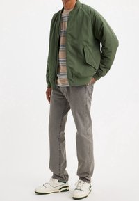 Hombre llevando una chaqueta bomber verde oliva, camisa de rayas, jeans grises y zapatillas blancas con acentos verdes, de pie contra un fondo blanco.
