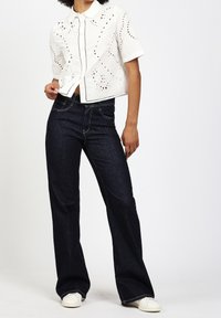 Camiseta blanca recortada con patrones cortados, combinada con jeans acampanados de color azul oscuro. El modelo lleva zapatillas blancas, con las manos en las caderas, contra un fondo liso.