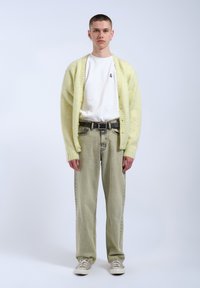 Ljusgul fluffig kofta, vit t-shirt och ljusgröna baggyjeans. En grå bälte och beige sneakers fulländar outfiten. En enfärgad bakgrund.