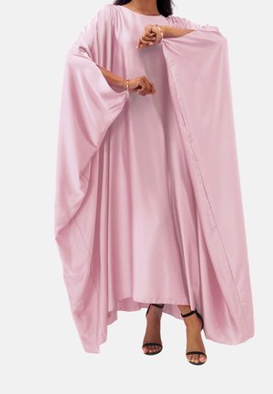 Femme portant une robe rose fluide avec de larges manches chauve-souris et des sandales noires à talons hauts, debout les bras levés.