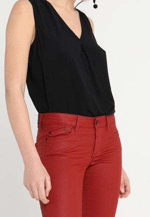 Vrouw draagt een mouwloze zwarte top met V-hals en aansluitende rode broek, waarbij het middelste deel van de romp en armen zichtbaar zijn tegen een effen witte achtergrond.