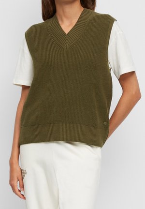 Persoon draagt een olijfgroen, gebreid mouwloos vest over een wit, kortärmelig shirt en witte broek, staand tegen een effen achtergrond.