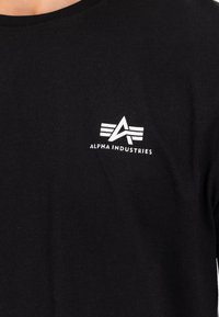 T-shirt de algodão preta com um logo branco de design angular no peito esquerdo, decote redondo e mangas curtas.