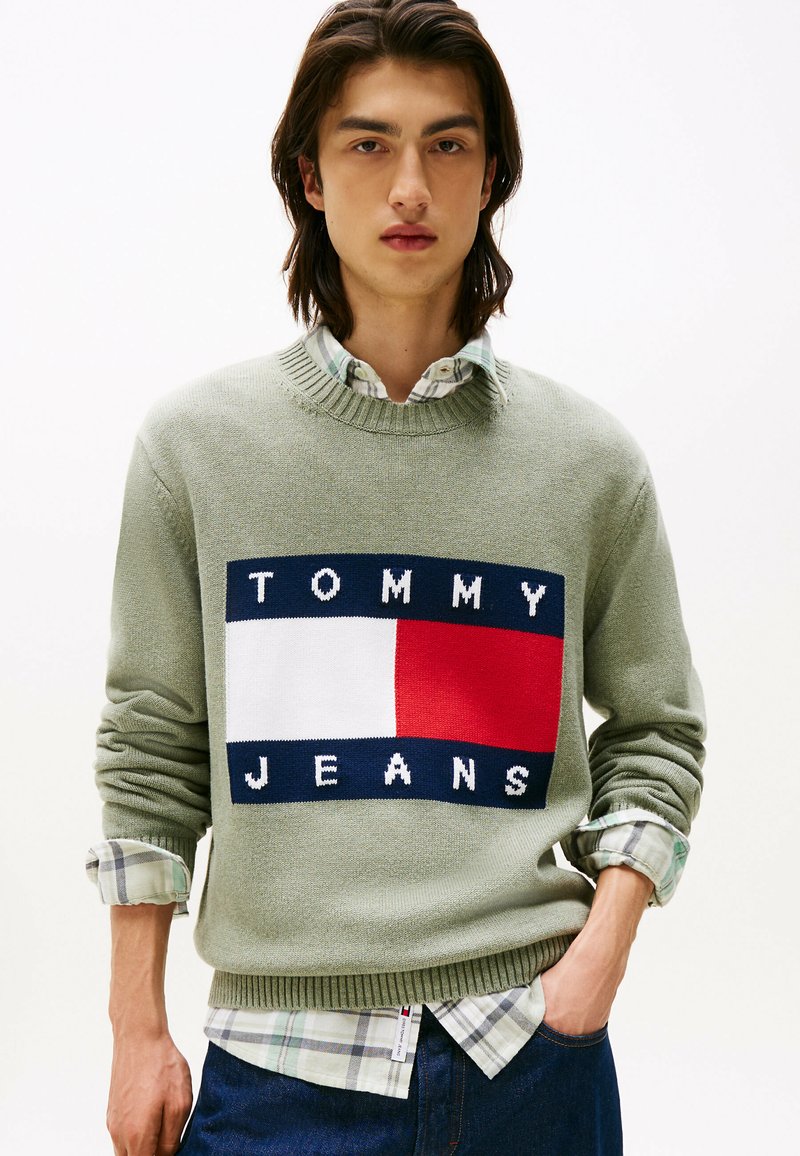 Maglione verde oliva con collo a coste, caratterizzato da un blocco logo blu navy, bianco e rosso che legge "TOMMY JEANS." Indossato sopra una camicia a scacchi.
