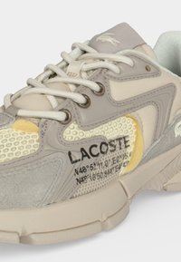 Μπεζ και γκρι αθλητικό παπούτσι Lacoste με λευκά κορδόνια, πάνελ από δίχτυ και σουέντ, με το όνομα της μάρκας και συντεταγμένες τυπωμένα στο πλάι.