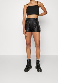 Top noir court avec des bretelles fines associé à un short en cuir noir taille haute avec des accents de boutons dorés et une finition texturée. Bottes de combat noires.