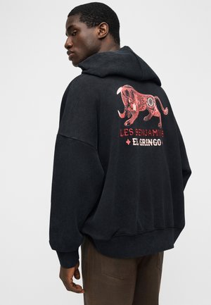 Άνδρας που φοράει μαύρη μπλούζα με κουκούλα με κόκκινο γραφικό μιας στυλιζαρισμένης ταύρου και το κείμενο «Les Benjamins El Gringo» στην πλάτη.