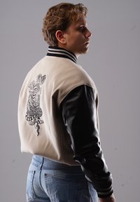 Cremfarbene und schwarze Collegejacke mit einem gestickten Tiger-Logo auf dem Rücken, geripptem Kragen und Ledermanschetten. Darunter sind Jeans aus Denim sichtbar.