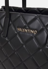 Svart vadderad läderväska med framträdande gyllene "Valentino"-logotyp, med eleganta handtag och en texturerad yta.