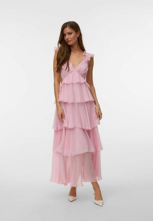 Femeie purtând o rochie maxi roz în straturi, fără mâneci, cu volane și decolteu în V, asortată cu pantofi albi cu vârf ascuțit, stând pe un fundal alb.