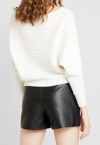 Pull blanc côtelé avec une coupe ample et des épaules tombantes, associé à un short en cuir noir, mettant en valeur une texture lisse et un design minimaliste.