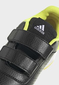 Zapato negro de cuero con detalles amarillos, correas de Velcro, lengüeta de malla, lados perforados y logotipo prominente de Adidas en el talón.