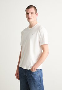 Witte katoenen t-shirt met korte mouwen, ronde hals en een subtiel grijs logo op de borst. Gecombineerd met blauwe denim jeans.