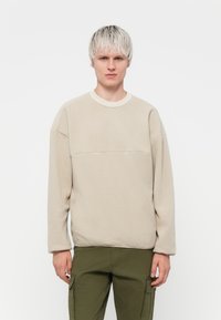 Sweatshirt en polaire beige avec un col rond, des manches longues et un détail de couture horizontal sur la poitrine. Texture douce, design minimaliste.