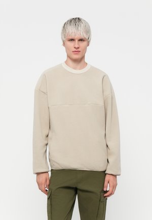 Beige fleece sweatshirt med rund halsudskæring, lange ærmer og en vandret sømdetalje over brystet. Blød tekstur, minimalistisk design.