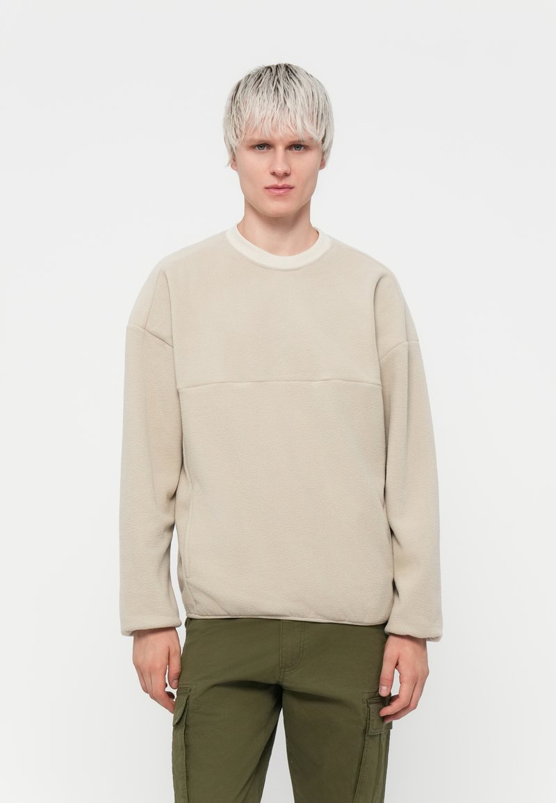 Sweatshirt en polaire beige avec un col rond, des manches longues et un détail de couture horizontal sur la poitrine. Texture douce, design minimaliste.
