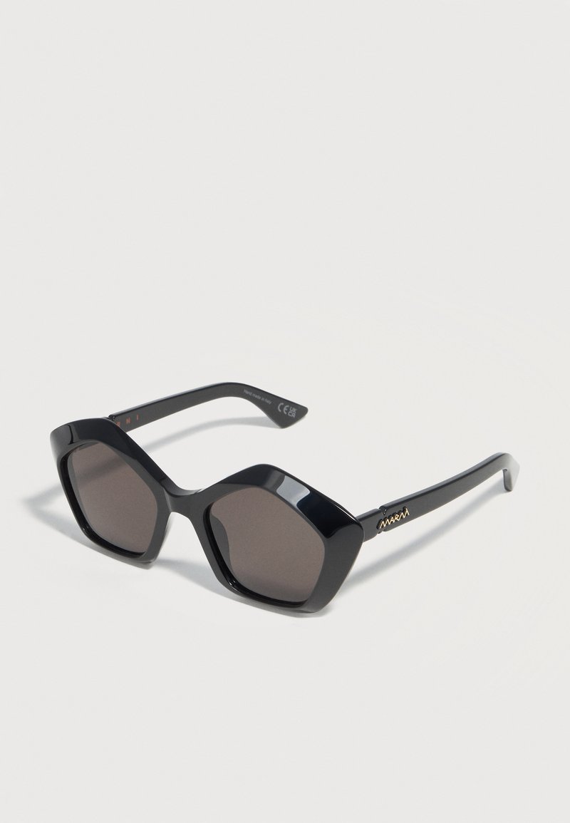 Black oversized hexagonal sunglasses with dark lenses and gold logo on the temple, displayed on a white surface.