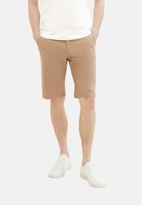 Homme portant un short beige mi-long, des baskets blanches et une chemise blanche, debout les mains dans les poches sur un fond uni.