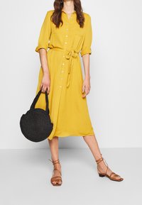 Abito giallo con bottoni, maniche corte, cintura da annodare e due tasche sul petto. Abbinato a sandali marroni con cinturini e a una borsa nera intrecciata.