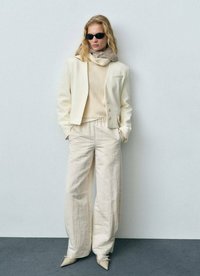 Blazer i krämfärg och beige stickad tröja, vida beige byxor och beige skor med spetsig tå. Modellen bär svarta solglasögon och en ljus scarf.
