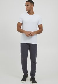 White short-sleeve t-shirt, round-neck design paired with dark gray joggers. Model wears black sneakers, standing with hands clasped.