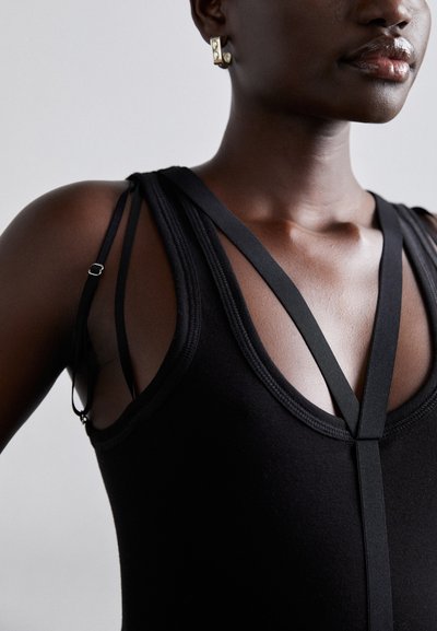 Fekete ujjatlan overall, vékonyan állítható pántokkal, mély V-nyakkivágással és sima, texturált anyaggal, melynek minimalista varrásai vannak.