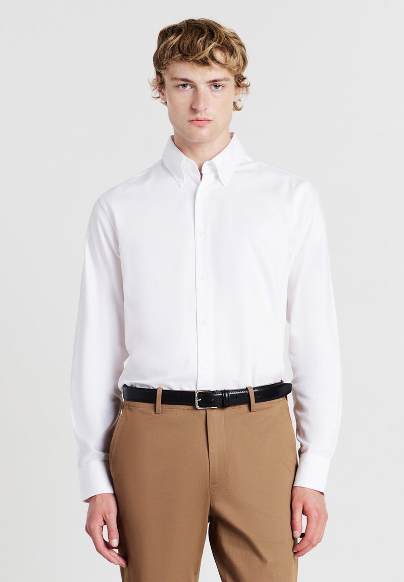 Camicia bianca a maniche lunghe con colletto a punta, chiusura con bottoni e una texture liscia, abbinata a pantaloni color sabbia e una cintura nera.