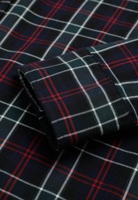 Camisa de cuadros oscura que presenta un patrón de cuadrícula en azul marino, blanco y rojo. La textura de la tela es suave, con un puño enrollado que muestra líneas definidas.