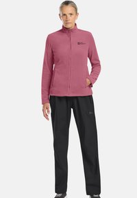 Giacca in pile rosa con chiusura a zip e due tasche laterali, abbinata a pantaloni neri con vita elastica e dettaglio del logo sulla gamba.