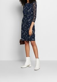Marineblaues Kleid mit Blumenmuster, langen transparenten Ärmeln, figurbetonter Silhouette und knielangem Saum, kombiniert mit weißen Stiefeletten.