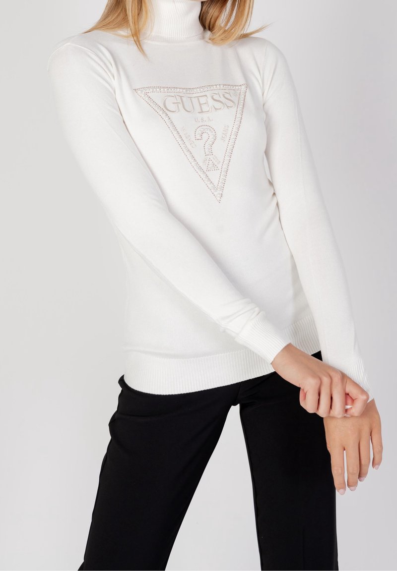 Femme portant un pull col roulé blanc Guess avec un logo en perles et un pantalon noir, posant avec les bras croisés aux poignets devant.