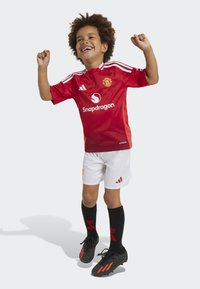 Maillot rouge de Manchester United avec un short blanc, accentué par des chaussettes noires et des chaussures noires avec des rayures orange. Enfant souriant en mouvement.